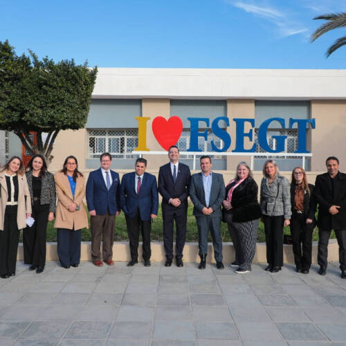 GVSU officials dressed in business and professional attire stands in a line outside in front of a building with a large sign that reads "I ♥ FSEGT." The scene features clear blue skies, a palm tree, and a trimmed tree.